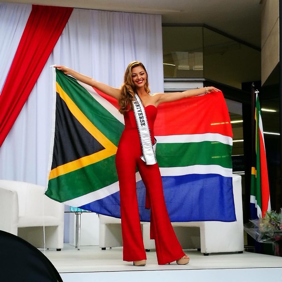 Pageant finalist in a white jumpsuit holding the South African flag